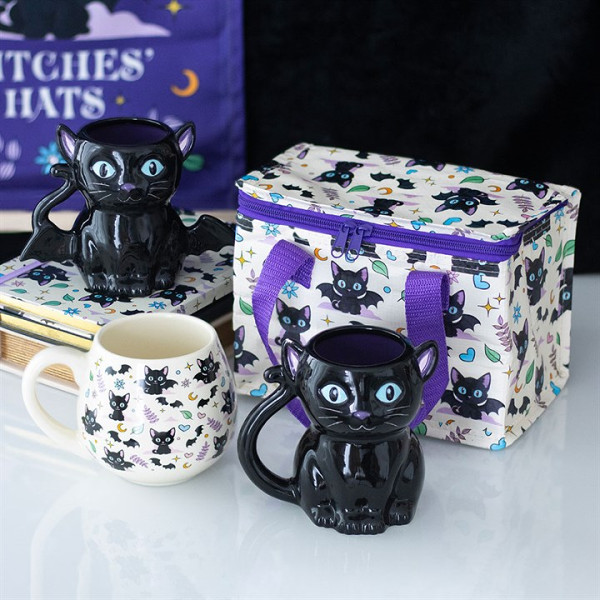 Black cat-themed mugs and a lunch bag with cat designs on a white surface.